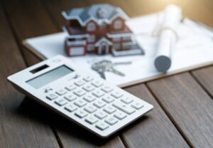 Calculadora junto a una maqueta de vivienda, llaves y planos, representando el cálculo fiscal de la exención por reinversión en vivienda habitual en el IRPF.