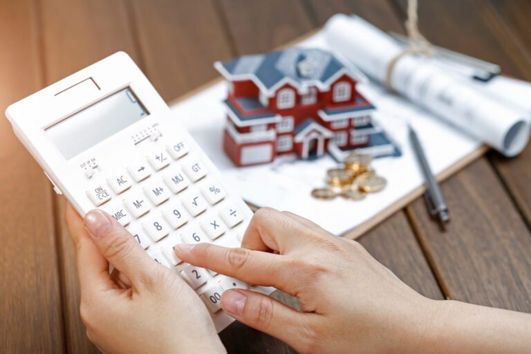 Mujer calculando la comisión de apertura hipoteca con una calculadora junto a una maqueta de casa y monedas